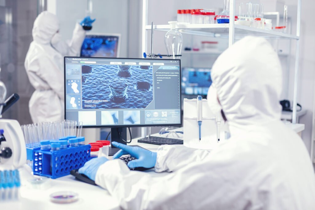 Chemist sits at his workplace in laboratory analyze coronavirus on computer dressed in ppe. Lab engineers conducting experiment for vaccine development against covid19 virus